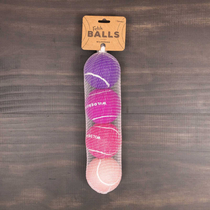 A mesh bag with three colorful Fetch Balls—purple, pink, and red—and a “Fetch BALLS WILDE DOG” cardboard label sits on a dark wooden surface.